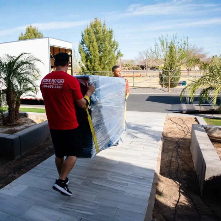 Midas Moving crew carrying a large wrapped furniture item along a home's front walkway toward a moving truck