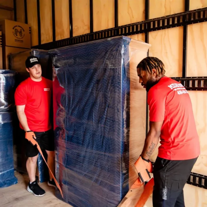 Two Midas Moving crew members using lifting straps to position a large wrapped cabinet inside a moving truck